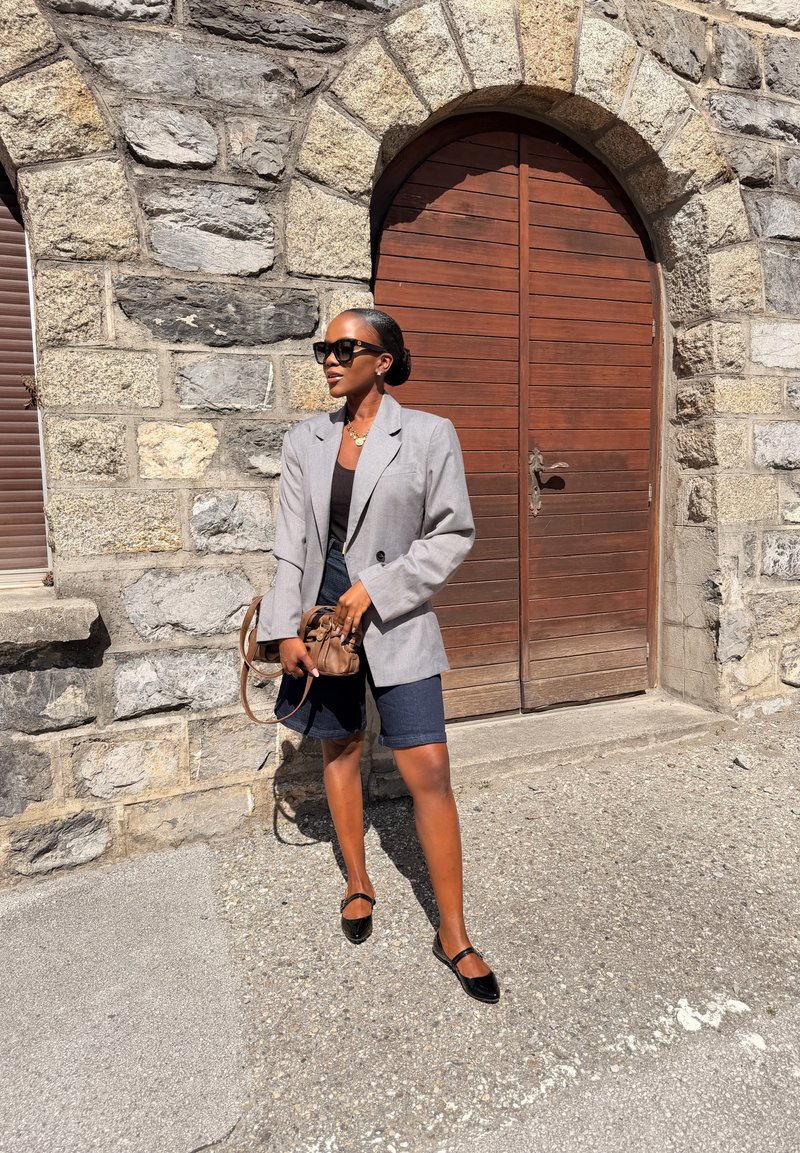 Donna con occhiali da sole, blazer grigio, top nero, shorts blu scuro e ballerine nere, in piedi con una borsa marrone vicino a un muro di pietra e una porta di legno.