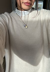 Pull en crème oversized avec un col en côte porté sur un turtleneck blanc. Deux colliers superposés avec des pendentifs en forme de cœur et rond.