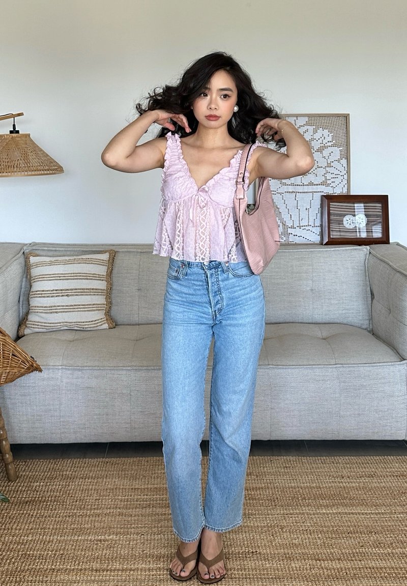 Young woman adjusting hair wearing a pink lace top, blue jeans, brown sandals, and carrying a pink shoulder bag in a modern living room.