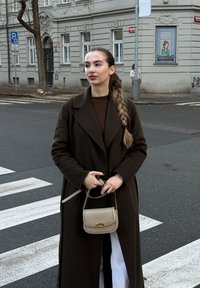 Long manteau brun foncé, pull marron, jupe blanche et petit sac bandoulière beige. Cheveux nattés, debout sur un passage piéton avec des bâtiments en arrière-plan.