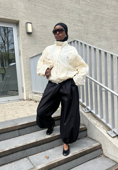 Mujer con chaqueta blanca de gran tamaño, pantalones anchos negros, pañuelo negro en la cabeza, gafas de sol, sosteniendo un bolso beige, de pie en unas escaleras exteriores junto a una barandilla.