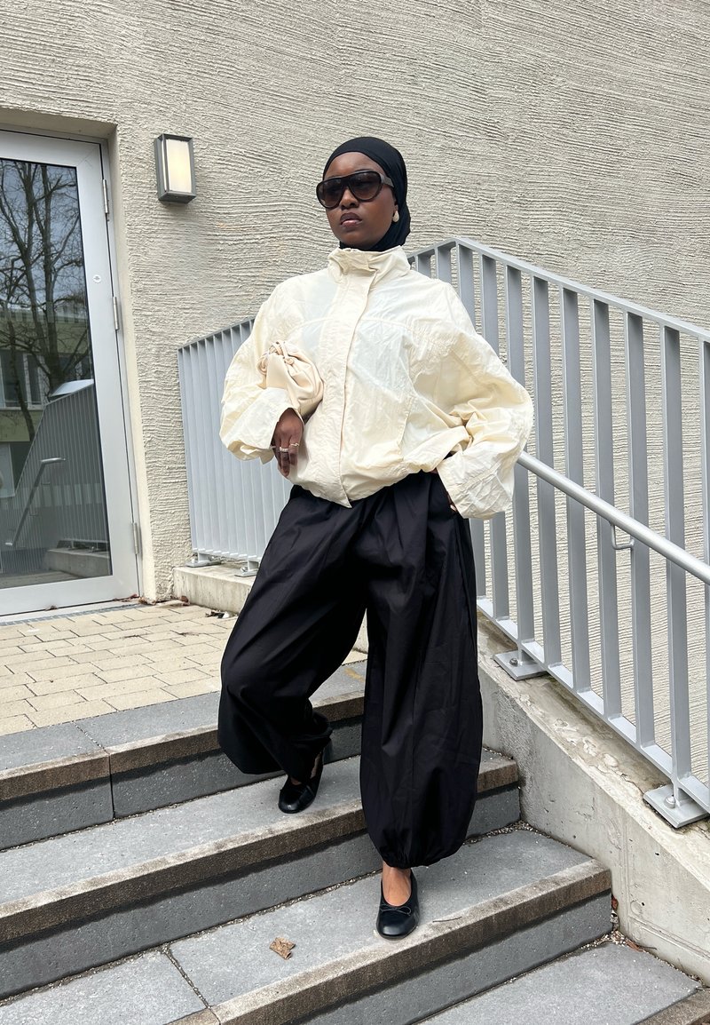 Sieviete valkā pārlielu baltu jaku, melnas platas bikses, melnu galvas lakatu, saules brilles, tur rokās bēša rokassomiņa, stāv ārējās kāpnēs pie margām.