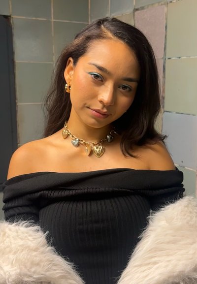 Top negro de canalé con hombros al descubierto, collar con colgante en forma de corazón dorado, pendientes de aro, suaves acentos de piel blanca. El fondo presenta una pared de azulejos.