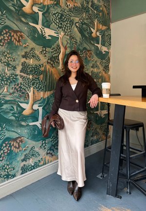 Mujer con gafas sonriente junto a una mesa de madera con una taza de café, frente a un mural con temática de naturaleza en tonos verdes y marrones.