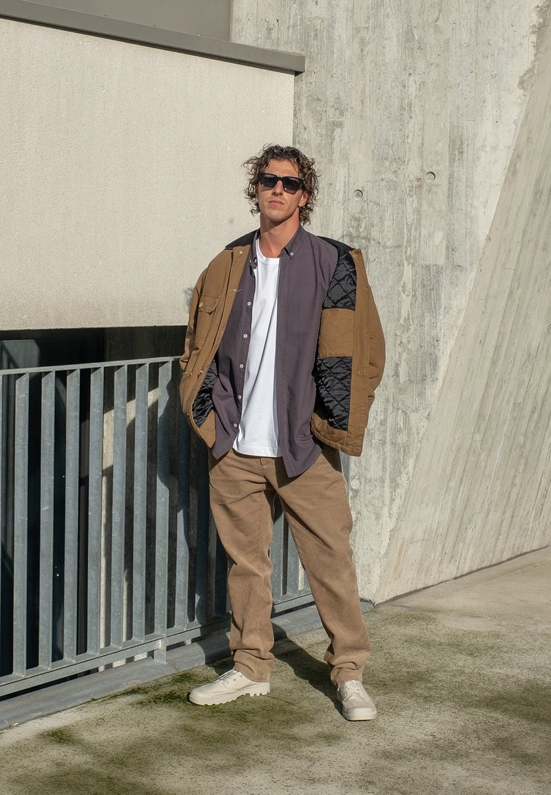 Burgundiškai siuvinėtas rudas švarkas virš pilko marškinėlių, derinamas su smėlio spalvos korduro linijomis ir šviesiais sportiniais bateliais. Vaizdą užbaigia akiniai nuo saulės.