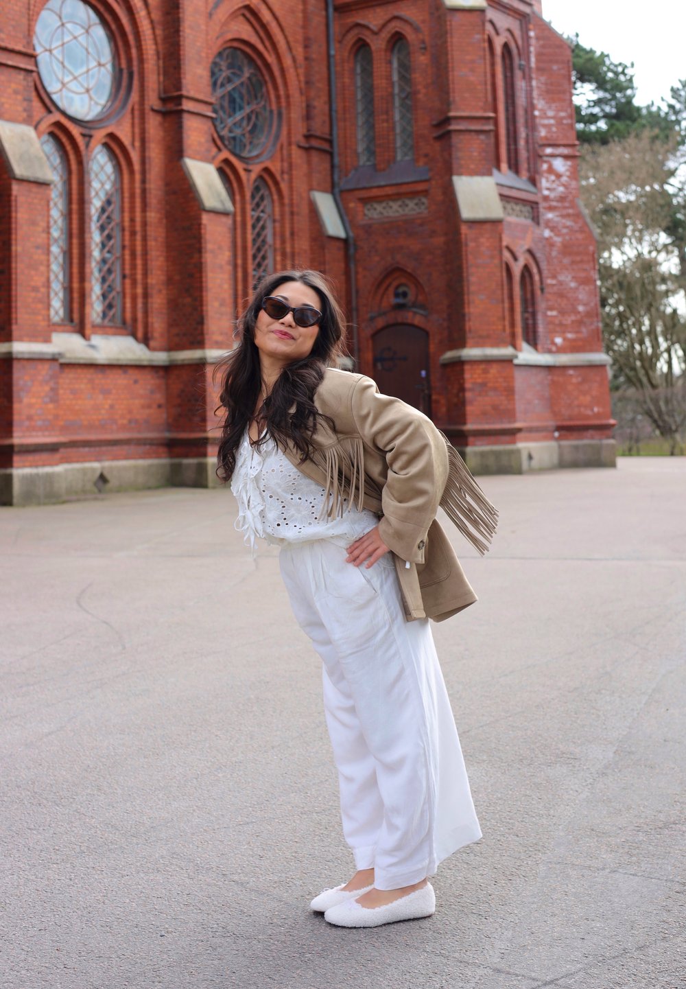 Brūnas frizūru jaka pār baltu mežģīņu topu un baltām platām biksēm, apvienota ar baltām pūkainām kurpēm, fona veido teksturēta sarkana ķieģeļu ēka.