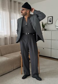 Veste de costume grise portée sur un henley gris foncé, avec un pantalon taille haute gris et des chaussures noires. Les accessoires comprennent un bonnet et un collier.