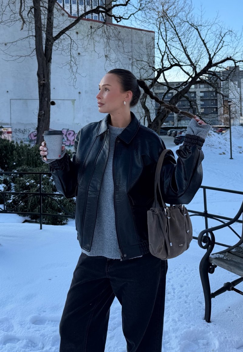 Mujer con chaqueta de cuero negra sosteniendo una taza de café y con el cabello trenzado, parada en un parque urbano nevado con edificios y árboles desnudos al fondo.