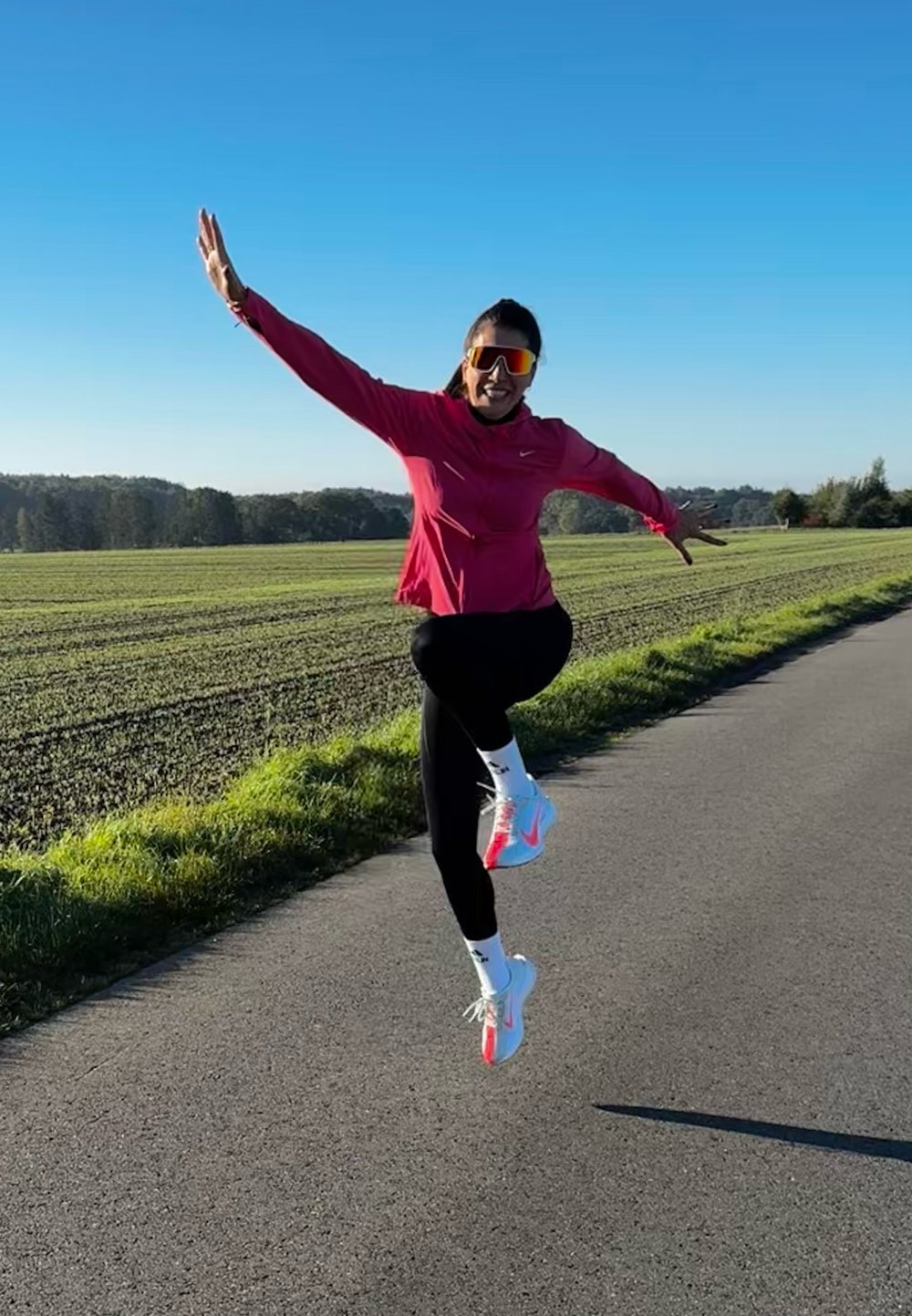 Sportlane, kes kannab roosat jakki ja musti retuuse, hüppab. Spordijalanõud on valged oranžide aktsentidega. Taustal on hele taevas ja roheline põld.
