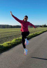 Sportlane, kes kannab roosat jakki ja musti retuuse, hüppab. Spordijalanõud on valged oranžide aktsentidega. Taustal on hele taevas ja roheline põld.