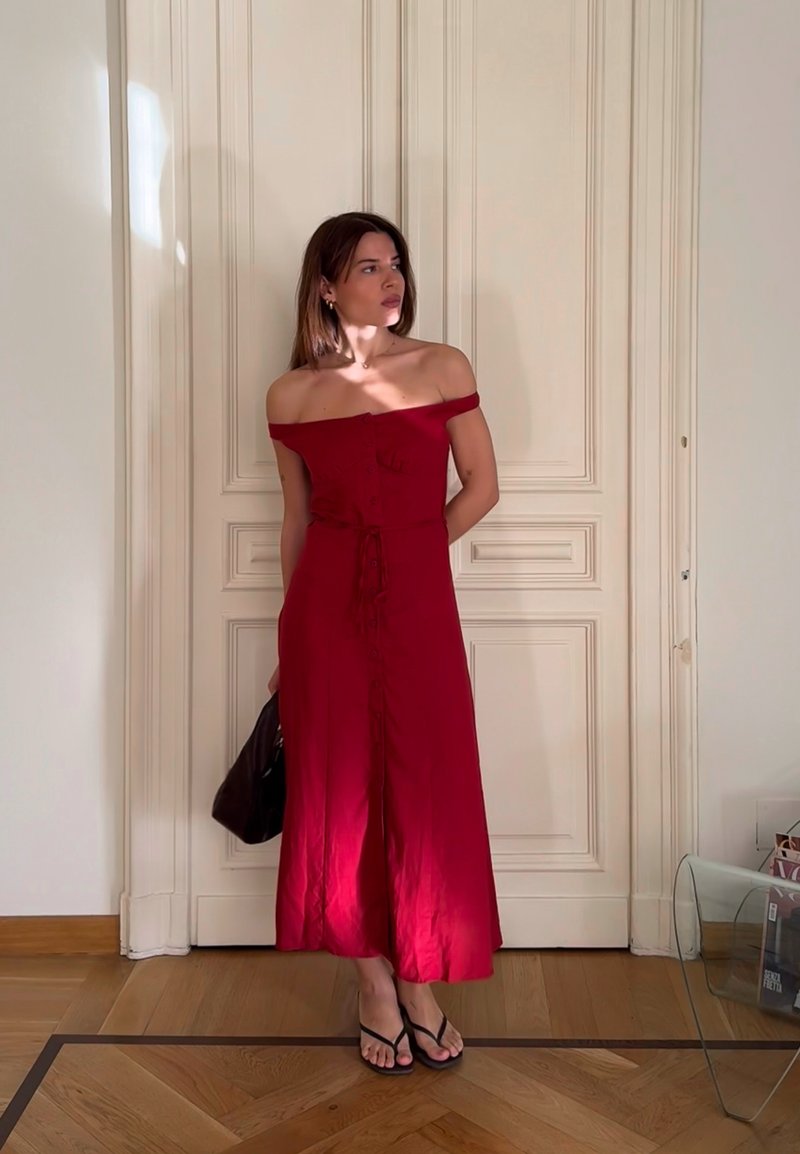 Vestido vermelho ombro a ombro com corpete justo, botões à frente, cinto atado na cintura e saia fluida. Usado com chinelos pretos e uma mala escura.