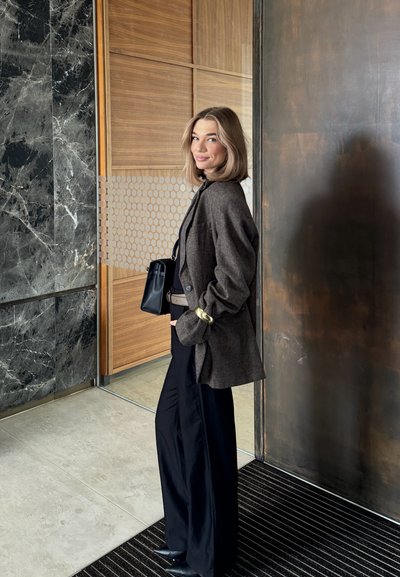 Blazer marrón oversize con textura de punto, pantalones negros de pierna ancha y botas negras puntiagudas. Hay un bolso negro y un acento dorado en la muñeca visibles.