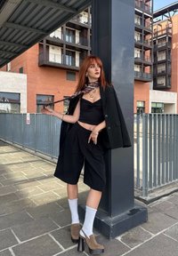 Crni oversize blejzer preko crnog crop topa i krojenih crnih šorts. Dodaci uključuju šal s uzorkom i debele smeđe cipele na petu.