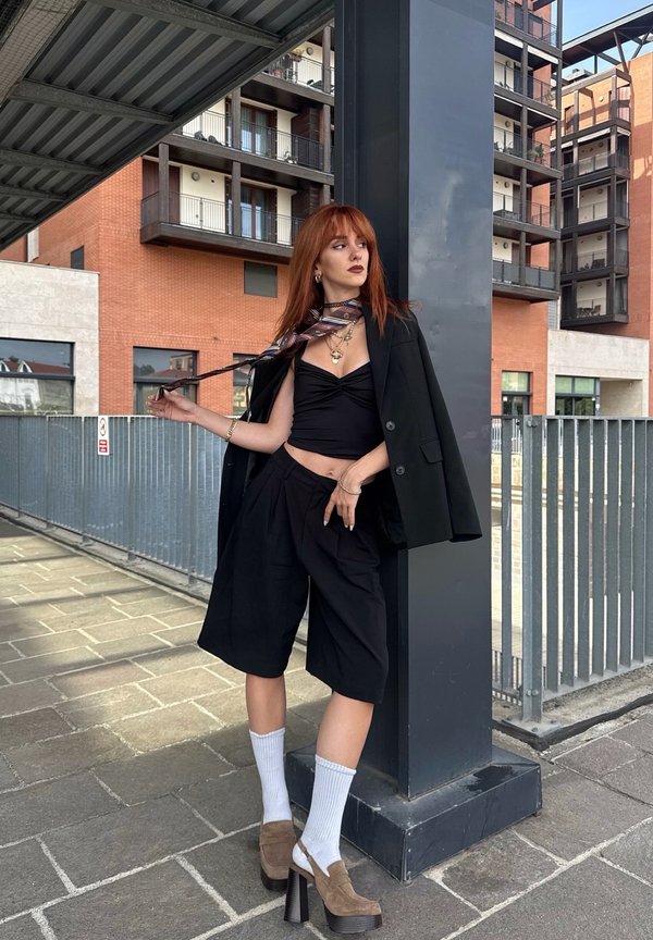 Zwarte oversized blazer gelaagd over een zwart crop top en op maat gemaakte zwarte shorts. Accessoires omvatten een gestreepte sjaal en stevige bruine hakken.
