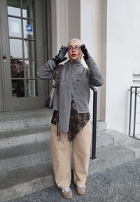 Femme portant un foulard beige, des lunettes, un pull gris boutonné, un haut en dentelle noire, un pantalon beige, des gants noirs et des baskets, ajustant ses lunettes devant un bâtiment.