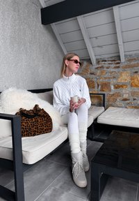 Femme en tricot blanc et bottes, assise sur un canapé beige tenant une tasse, avec un sac à imprimé léopard et un mur en pierre en arrière-plan.