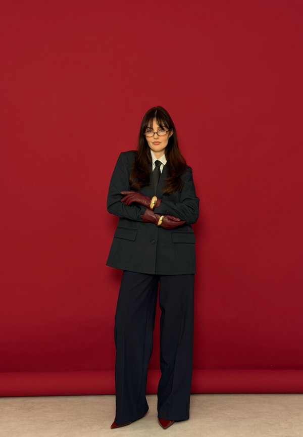 Žena v čiernom obleku, bielej košeli, čiernej kravate, bordových rukaviciach a okuliaroch stojí so založenými rukami pred červeným pozadím.