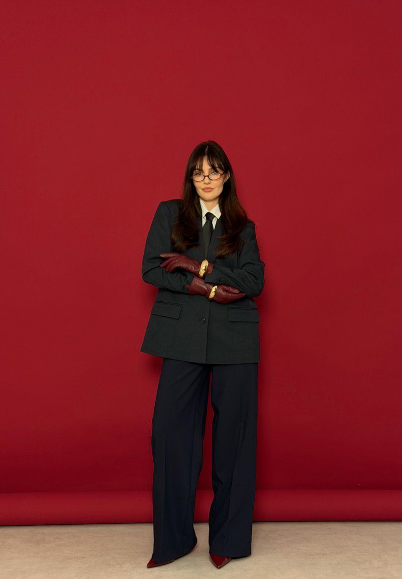 Donna con abito nero, camicia bianca, cravatta nera, guanti color borgogna e occhiali, in piedi con le braccia incrociate davanti a uno sfondo rosso.