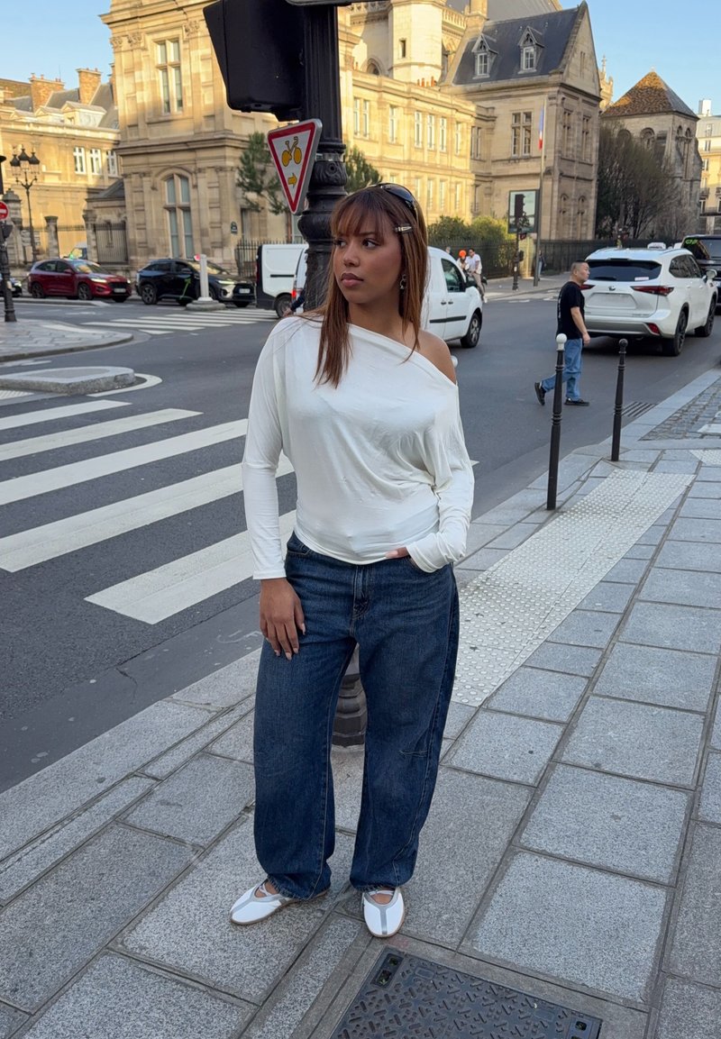Νέα γυναίκα με λευκό τοπ που αφήνει τους ώμους ακάλυπτους και μπλε τζιν στέκεται σε πεζοδρόμιο της πόλης κοντά σε διάβαση πεζών και ιστορικά κτίρια.