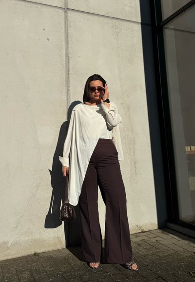 Mujer con gafas de sol y una camiseta blanca asimétrica con pantalones anchos oscuros, sosteniendo un bolso marrón contra una pared de hormigón.