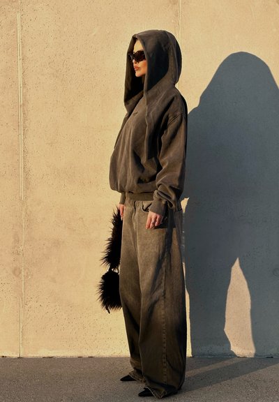 Sudadera de algodón gris oscuro de gran tamaño con capucha; pantalones grises de corte holgado; sostiene un accesorio de piel negra; de pie junto a una pared de concreto.