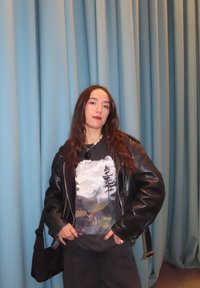 Čierna kožená bunda cez tričko s grafikou horskej scenérie, tmavé nohavice a kabelka cez rameno. Vlnité vlasy, stojí pred svetlomodrými závesmi.