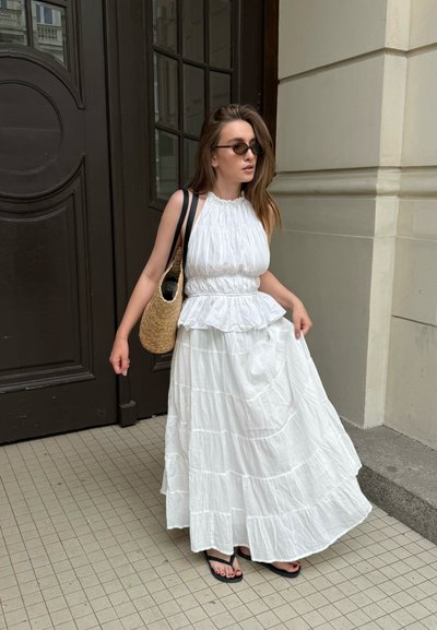 Vestido maxi blanco con capas y dobladillo rizado, cuerpo recogido y escote halter. Combinado con un bolso de paja tejida y chanclas negras.
