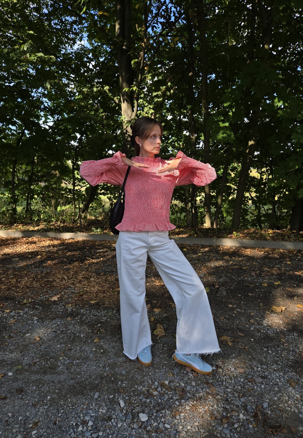 Rote und weiße gemusterte Bluse mit Glockenärmeln, kombiniert mit weißen weiten Hosen und blauen Turnschuhen, steht auf einer Kiesoberfläche.