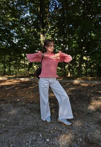 Rote und weiße gemusterte Bluse mit Glockenärmeln, kombiniert mit weißen weiten Hosen und blauen Turnschuhen, steht auf einer Kiesoberfläche.