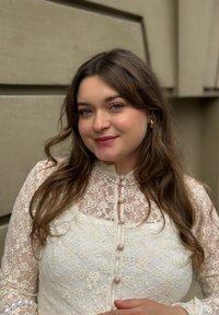 Ženska z dolgimi rjavimi lasmi se smeška, oblečena v belo čipkast zgornji del s perladnimi gumbi, stoji ob bež zunanjosti stene.
