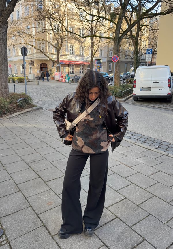 Schwarze Lederjacke, Pullover mit Tarnmuster in Grau und Braun, schwarze weit geschnittene Hose und schwarze Plateau-Schuhe. Straßenszenerie.
