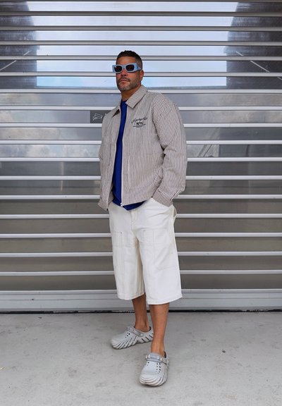 Chaqueta ligera a rayas, camisa azul, pantalones cortos cargo blancos, zuecos grises y gafas de sol azules; de pie contra un fondo de listones grises.