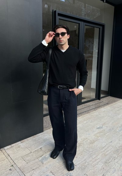 Hombre con suéter negro, pantalones anchos de color azul marino, zapatos negros de dedos partidos y gafas de sol, de pie frente a una puerta de cristal con una bolsa negra al hombro.