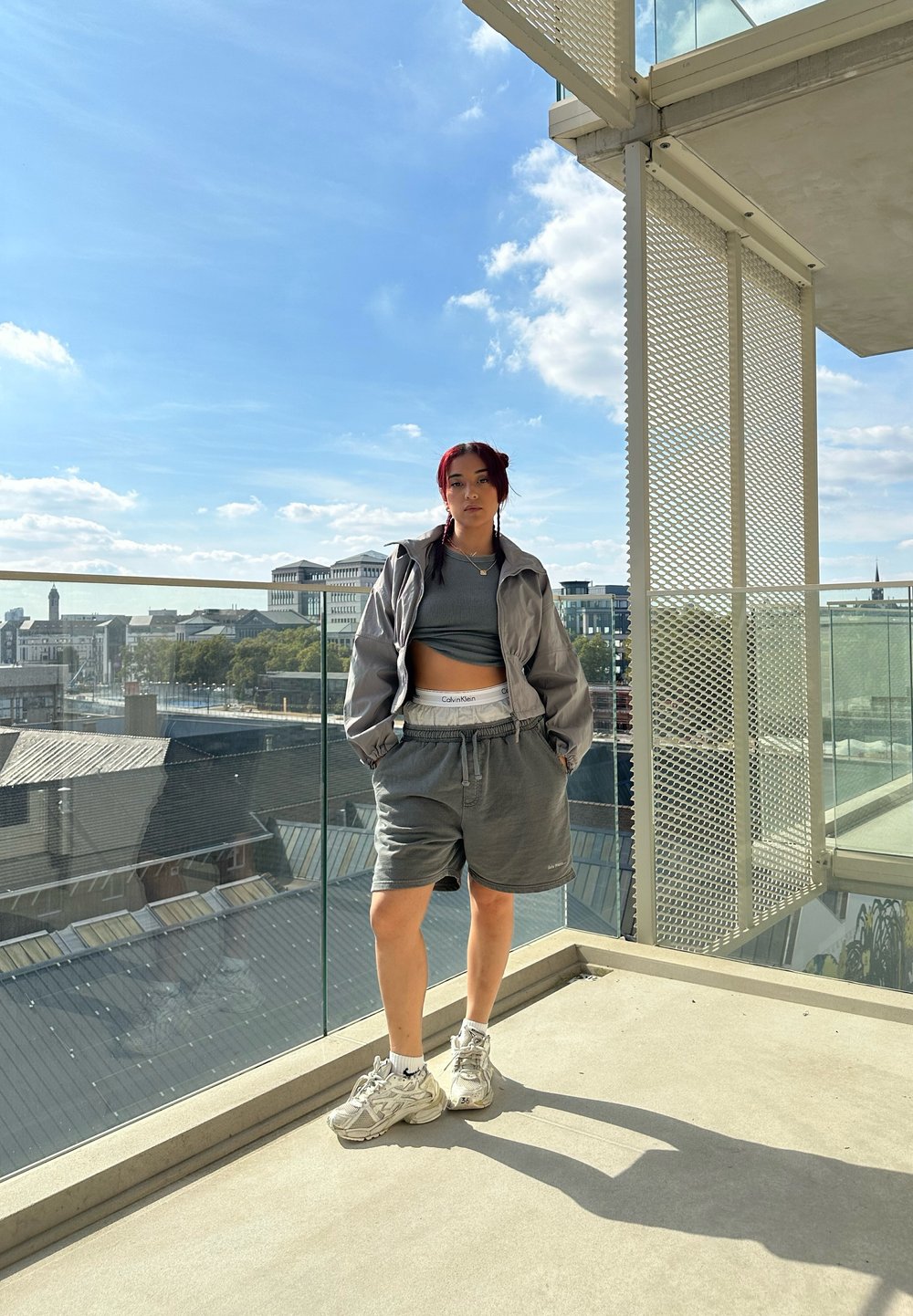 Grijze cropped jas over een grijze tanktop en shorts, gecombineerd met sneakers. Buitengevel met glazen reling, blauwe lucht en wolken.