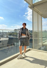 Grijze cropped jas over een grijze tanktop en shorts, gecombineerd met sneakers. Buitengevel met glazen reling, blauwe lucht en wolken.