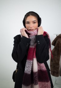 Villahuivi punaisilla, vaaleanpunaisilla ja harmailla ruudullisilla kuvioilla, ripustettuna kaulan ympäri. Musta takki ja tekoturkiskuulokkeet täydentävät ilmeen.