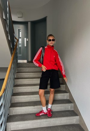 Veste zippée rouge avec des rayures blanches, shorts en denim noir, baskets rouges, et sac à pois noirs sur un escalier avec des murs gris et une balustrade.