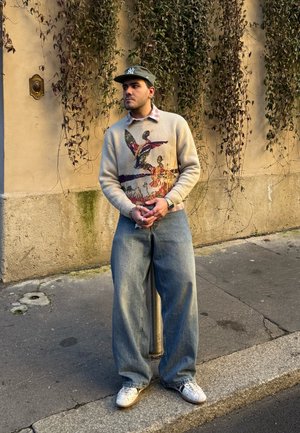 Joven con un suéter gráfico, jeans anchos, zapatillas blancas y una gorra verde, de pie en una acera frente a una pared beige con enredaderas colgantes.