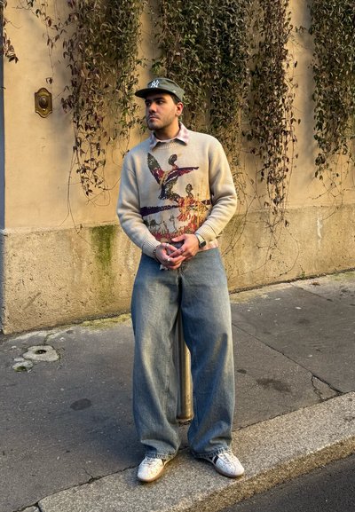 Joven con un suéter gráfico, jeans anchos, zapatillas blancas y una gorra verde, de pie en una acera frente a una pared beige con enredaderas colgantes.