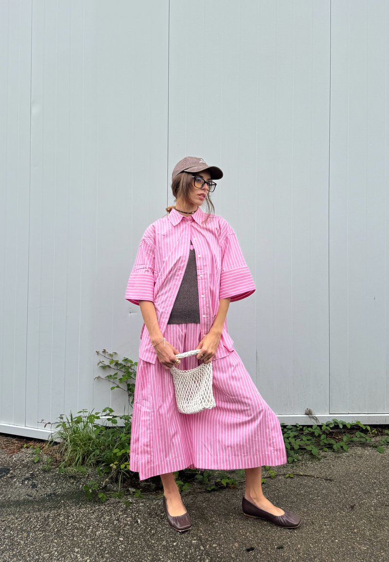 Camisa oversized às riscas cor-de-rosa com saia a condizer, blusa cinza por baixo, chapéu castanho, óculos e sapatilhas castanhas. A carregar um saco de palha.