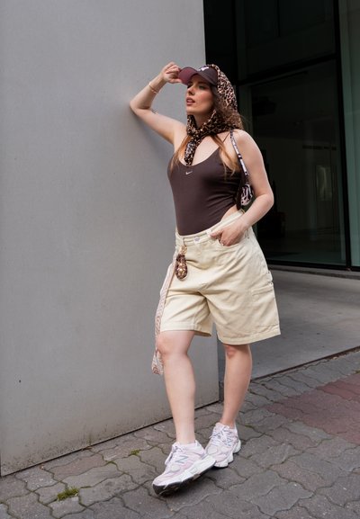 Mujer joven con una camiseta sin mangas marrón, pantalones cortos beige, zapatillas rosas, bufanda de estampado de leopardo y gorra, posando frente a una pared gris al aire libre.