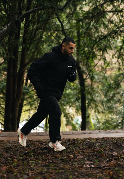 Hombre corriendo por un sendero forestal con ropa deportiva negra y zapatillas blancas de correr con detalles en naranja.