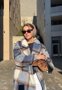 Femme debout en plein air entre des bâtiments, portant un manteau à carreaux, des cache-oreilles, des lunettes de soleil, tenant des sacs pliés marron et noir sous la lumière du soleil.