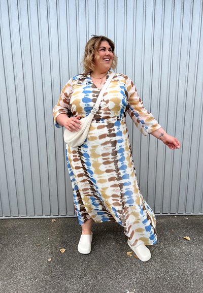 Vestido largo y fluido en un patrón de tie-dye en beige, azul y marrón. Tiene mangas de tres cuartos y cintura ceñida. Combinado con zuecos blancos.