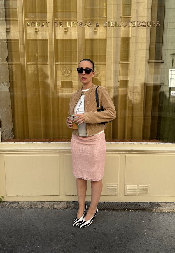 Femeie cu ochelari de soare stă în fața unei librării, poartă o jachetă bej, o cămașă albă, o fustă roz, pantofi cu model și ține o băutură în mână.