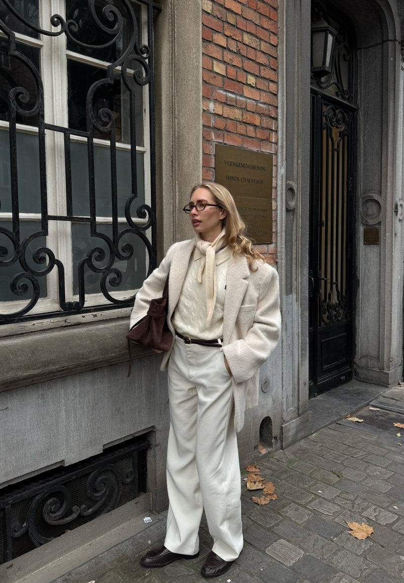 Cremefarbener übergroßer Wollmantel über einem beigefarbenen Stricktop, kombiniert mit weißen, weiten Hosen und braunen Loafers, dazu eine braune Handtasche.