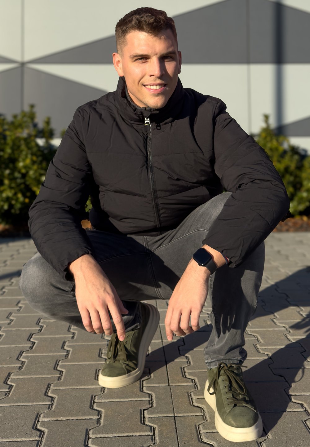 Μαύρο μπουφάν, γκρίζα τζιν, ελιόχρωμα αθλητικά παπούτσια με κρέμες σόλες. Το άτομο είναι σε στατικότητα σε μια διακοσμημένη επιφάνεια από πέτρα.