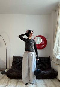 Haut noir à manches courtes avec un design texturé, associé à un pantalon de survêtement gris ample. La personne tient un petit sac noir, debout dans une pièce éclairée doucement.