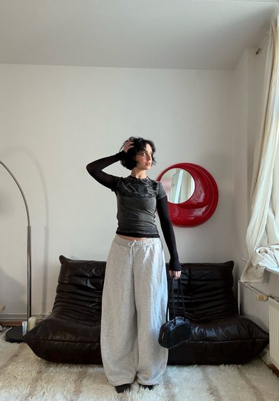 Top negro de manga corta con diseño texturizado, combinado con pantalones deportivos grises holgados. Persona sostiene una pequeña bolsa negra, de pie en una habitación con luz suave.