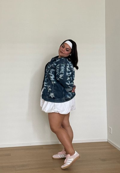 Chaqueta de denim con texto estilo graffiti, falda blanca, zapatillas rosas con tres franjas y una diadema blanca. Fondo neutro y suelo de madera.